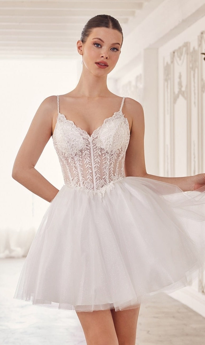 A woman stands indoors in a bright, elegant room wearing the Ladivine Embroidered Lace Homecoming Dress CD0236, featuring a short white design with a corset bodice and thin straps.